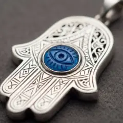 Ett silverhänge i form av hand of fatima med ett blått öga i mitten. Detaljerade mönster pryder hänget.