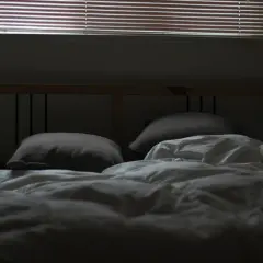 En obäddad säng med kuddar och vitt täcke, redo för dua sleep. Persiennerna släpper in svagt ljus.