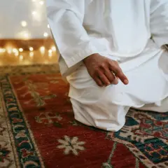 En person i vit klädnad sitter på en matta och ber. Detta är en del av vad som är salat, en viktig bön inom islam.
