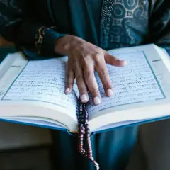 En hand vilar på en öppen Koran, med ett radband hängande över sidorna. En stilla stund för bön, kanske under fajr.