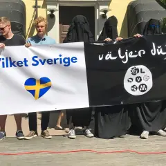 Människor håller en banderoll med texten "Vilket Sverige" och en svensk hjärta, samt "Väljer du?". Debatten om böneutrop i Sverige fortsätter.