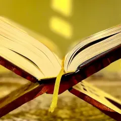 En öppen bok på ett träställ, med en gul bokmärkesflik. Kanske är det surah Naba som läses.