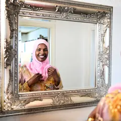 En kvinna i rosa hijab justerar sin klänning framför en utsmyckad spegel. Hon ler glatt, som om hon förbereder sig för ett speciellt tillfälle, kanske som Muhammeds fru Khadija en gång gjorde.