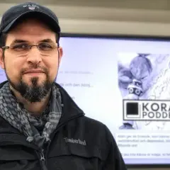 Man med skägg och keps ler mot kameran. I bakgrunden syns en skärm med texten "KORAN PODDEN" och arabisk skrift. Allahu alam.