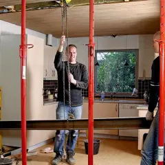 Två män arbetar med att säkra ett tak med stödpålar och en balk. En kedja hänger från taket, som om de förbereder för att lyfta något, kanske en del av surah balad.