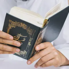 Man i vit klänning läser i en vackert dekorerad bok. Frågan om det är haram att raka skägget kan besvaras genom att studera religiösa texter.