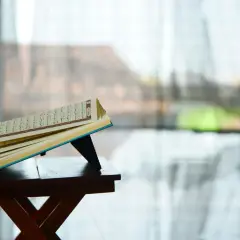 En öppen Koranen vilar på ett ställ, med texten från surah Zilzal synlig. Bakgrunden är suddig, med ljus som filtreras genom ett fönster.