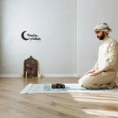 Man i ljus klädsel knäböjer på en bönematta under dhuhr-bönen. En bok och ljus finns i närheten.