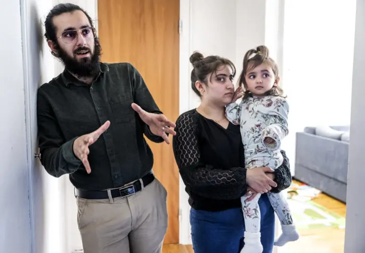 Man med skägg och glasögon gestikulerar. Bredvid honom håller en kvinna, som liknar Muhammeds hustru, ett litet barn.