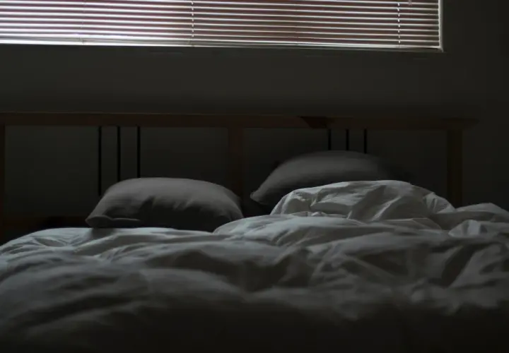 En obäddad säng med kuddar och vitt täcke, redo för dua sleep. Persiennerna släpper in svagt ljus.