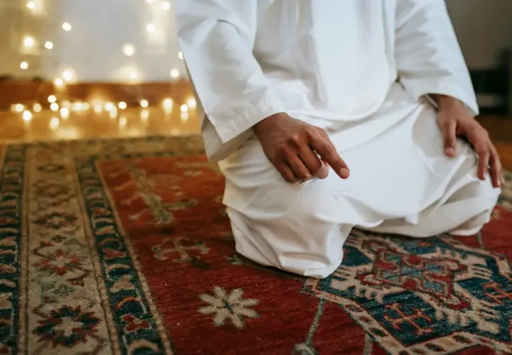 En person i vit klädnad sitter på en matta och ber. Detta är en del av vad som är salat, en viktig bön inom islam.