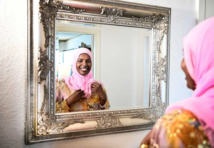 En kvinna i rosa hijab justerar sin klänning framför en utsmyckad spegel. Hon ler glatt, som om hon förbereder sig för ett speciellt tillfälle, kanske som Muhammeds fru Khadija en gång gjorde.