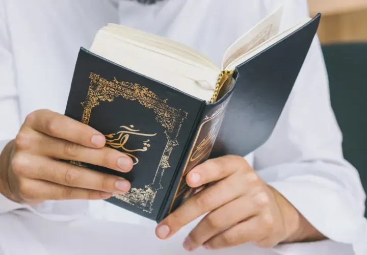 Man i vit klänning läser i en vackert dekorerad bok. Frågan om det är haram att raka skägget kan besvaras genom att studera religiösa texter.