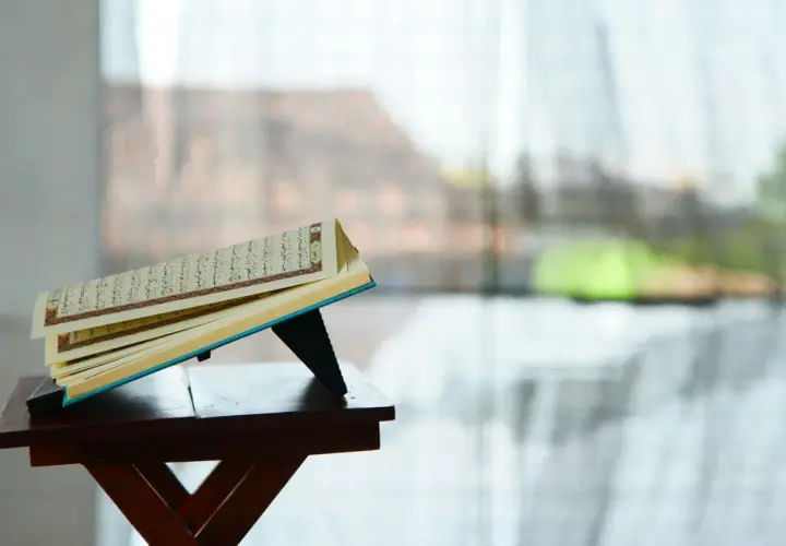 En öppen Koranen vilar på ett ställ, med texten från surah Zilzal synlig. Bakgrunden är suddig, med ljus som filtreras genom ett fönster.