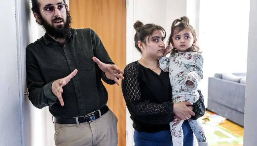 Man med skägg och glasögon gestikulerar. Bredvid honom håller en kvinna, som liknar Muhammeds hustru, ett litet barn.