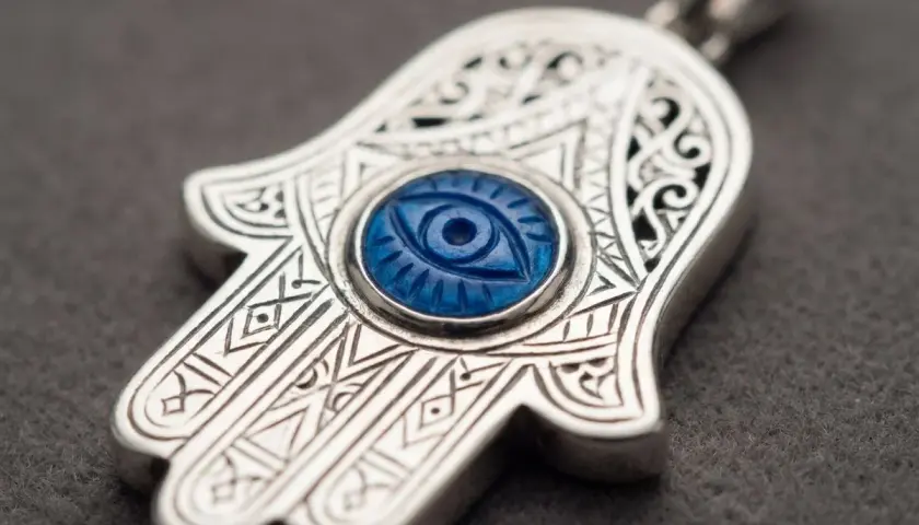 Ett silverhänge i form av hand of fatima med ett blått öga i mitten. Detaljerade mönster pryder hänget.
