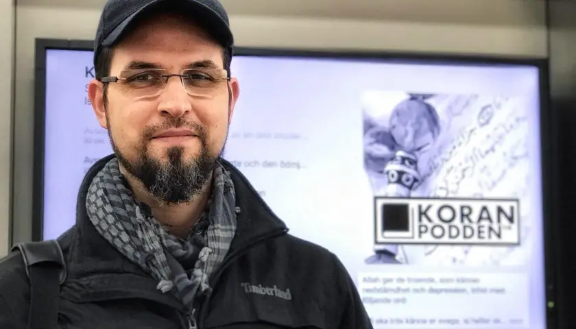Man med skägg och keps framför en skärm med texten "KORAN PODDEN". Han ser tacksam ut, som om han ber: "ربي اوزعني ان اشكر نعمتك".