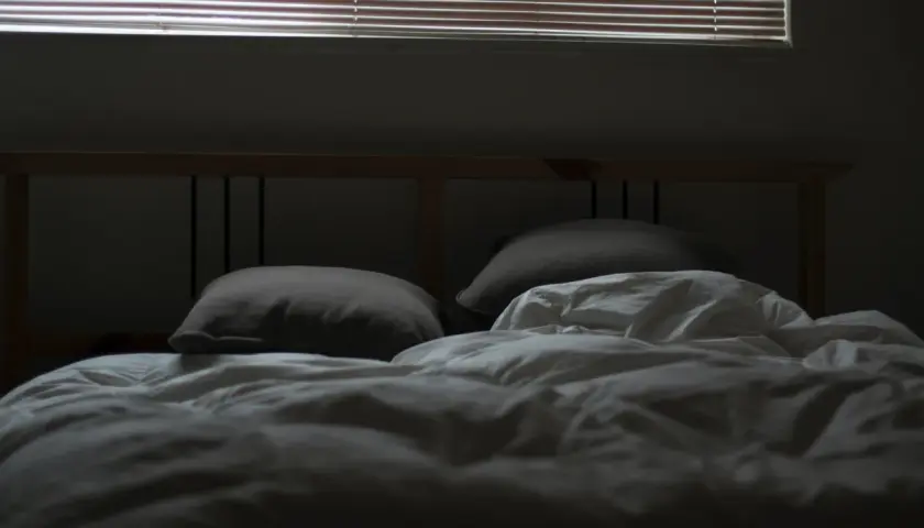 En obäddad säng med kuddar och vitt täcke, redo för dua sleep. Persiennerna släpper in svagt ljus.