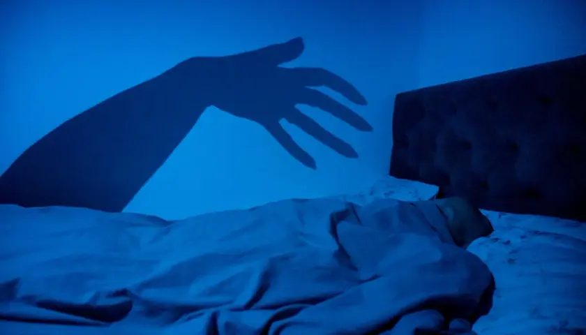 En skugga av en hand sträcker sig mot en sovande person i mörkret, en bild som kan påminna om upplevelser av sleep paralysis inom islam.
