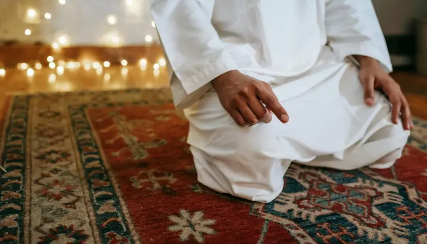En person i vit klädnad sitter på en matta och ber. Detta är en del av vad som är salat, en viktig bön inom islam.