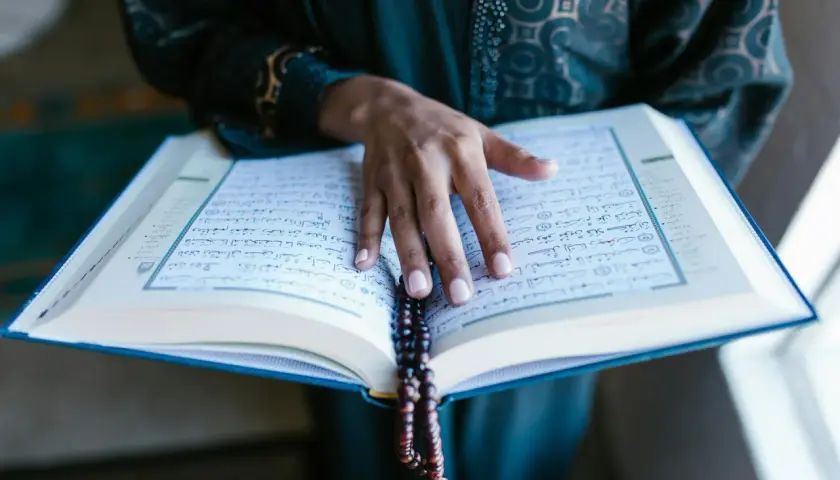 En hand vilar på en öppen Koran, med ett radband hängande över sidorna. En stilla stund för bön, kanske under fajr.