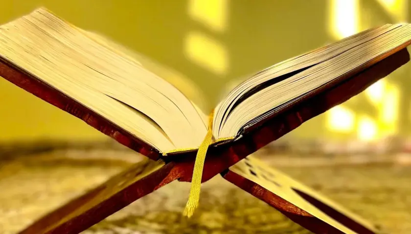 En öppen bok på ett träställ, med en gul bokmärkesflik. Kanske är det surah Naba som läses.