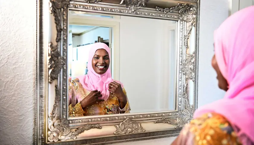 En kvinna i rosa hijab justerar sin klänning framför en utsmyckad spegel. Hon ler glatt, som om hon förbereder sig för ett speciellt tillfälle, kanske som Muhammeds fru Khadija en gång gjorde.