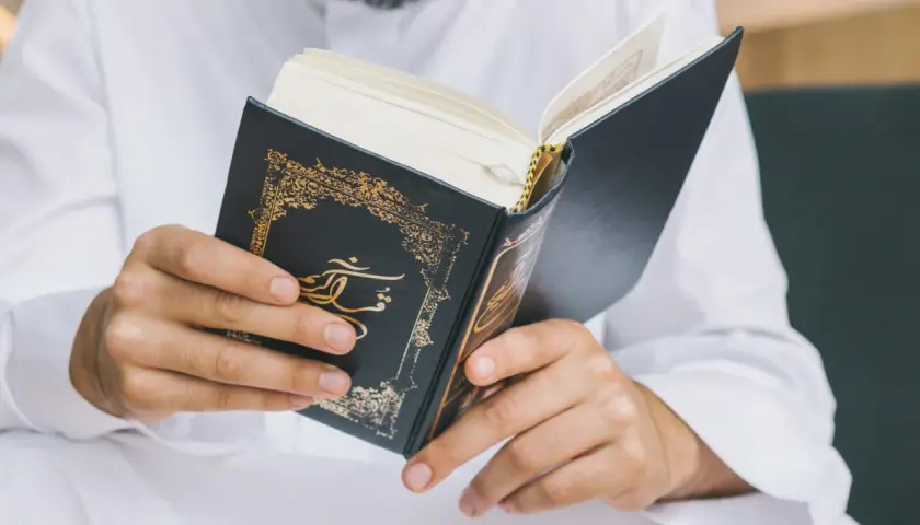 Man i vit klänning läser i en vackert dekorerad bok. Frågan om det är haram att raka skägget kan besvaras genom att studera religiösa texter.