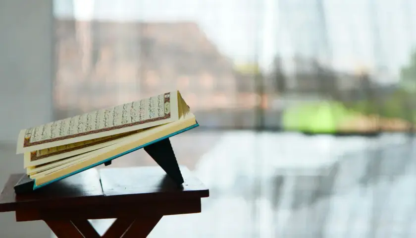 En öppen Koranen vilar på ett ställ, med texten från surah Zilzal synlig. Bakgrunden är suddig, med ljus som filtreras genom ett fönster.