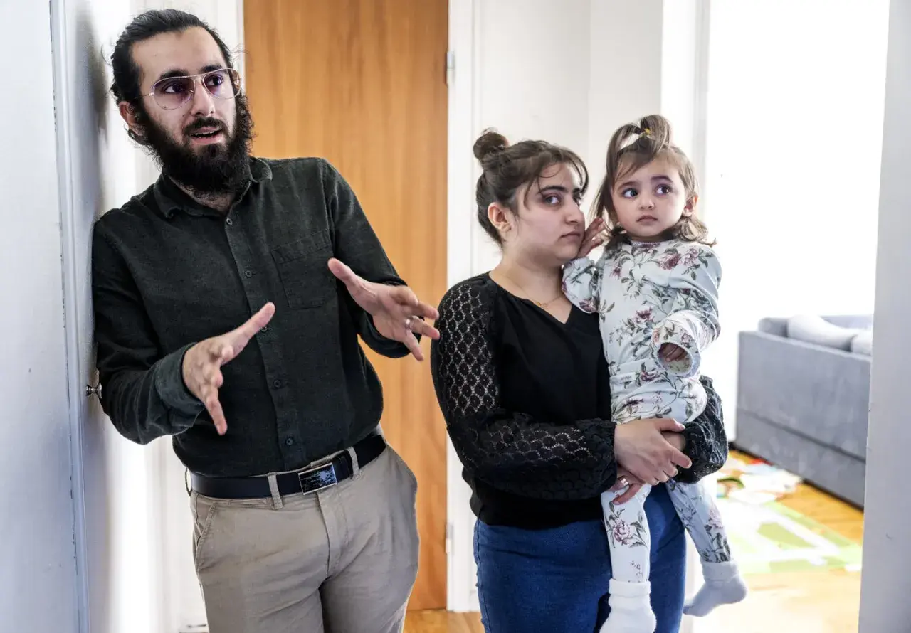 Man med skägg och glasögon gestikulerar. Bredvid honom håller en kvinna, som liknar Muhammeds hustru, ett litet barn.