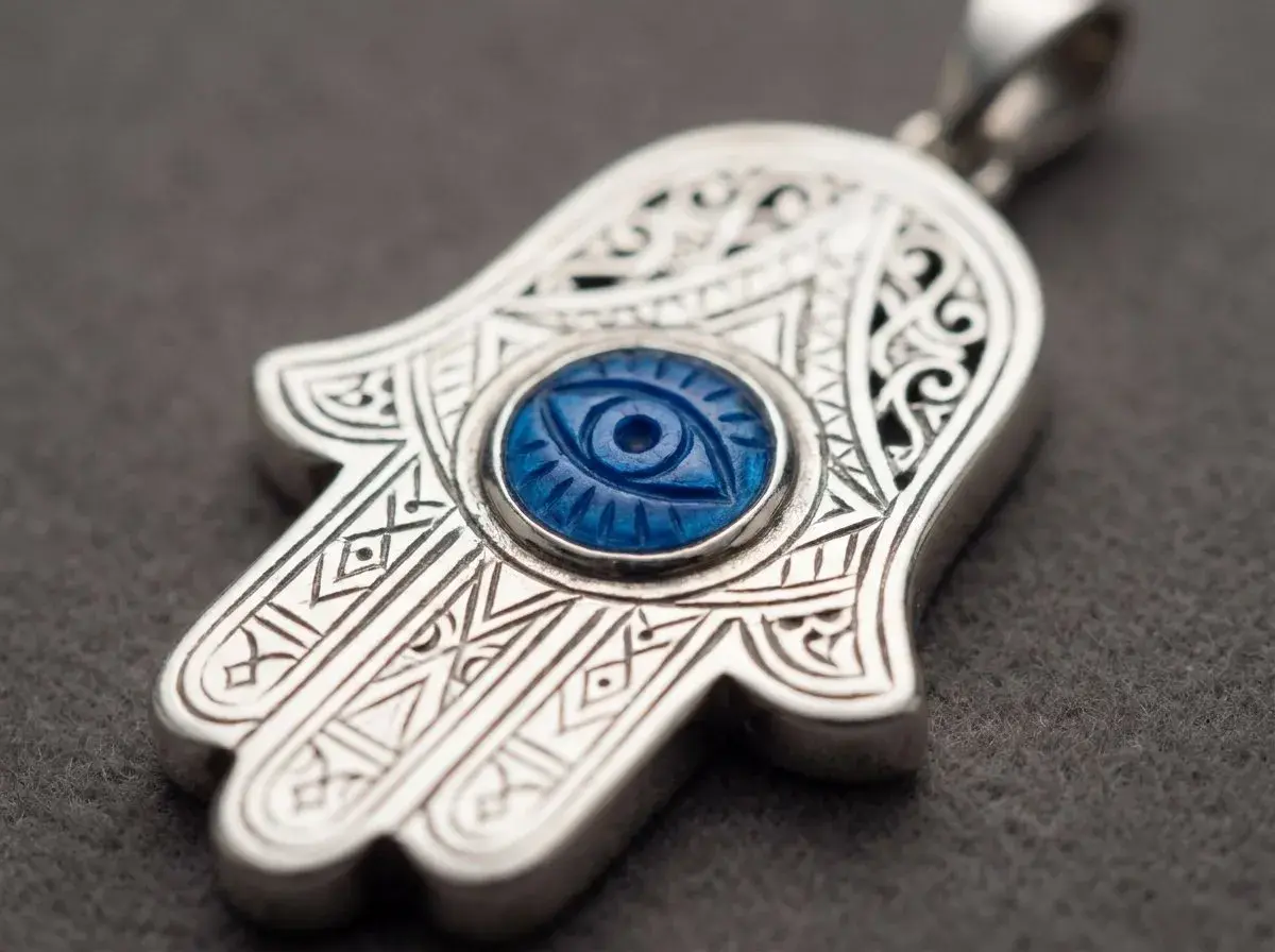 Ett silverhänge i form av hand of fatima med ett blått öga i mitten. Detaljerade mönster pryder hänget.
