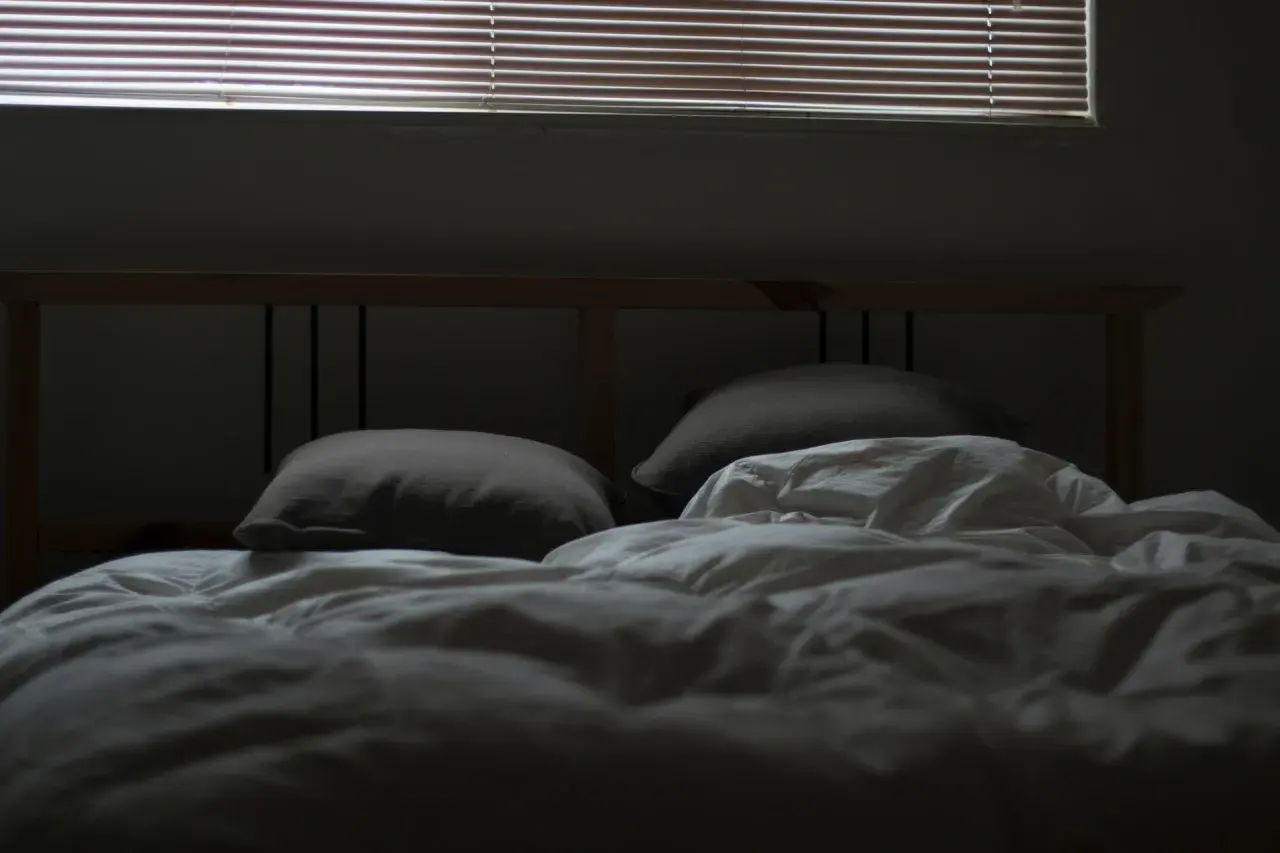 En obäddad säng med kuddar och vitt täcke, redo för dua sleep. Persiennerna släpper in svagt ljus.