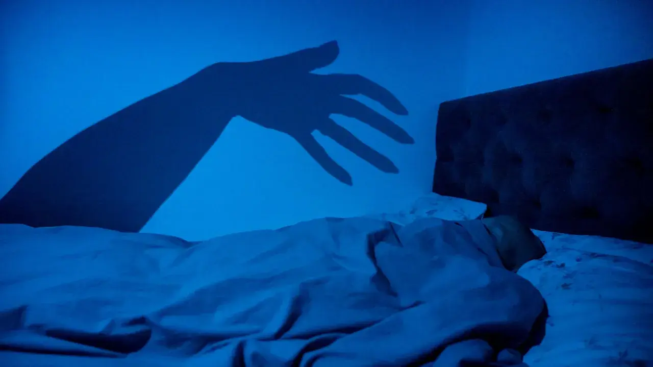 En skugga av en hand sträcker sig mot en sovande person i mörkret, en bild som kan påminna om upplevelser av sleep paralysis inom islam.