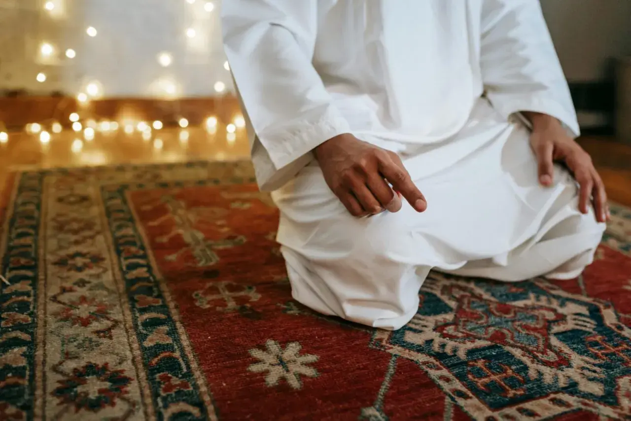 En person i vit klädnad sitter på en matta och ber. Detta är en del av vad som är salat, en viktig bön inom islam.