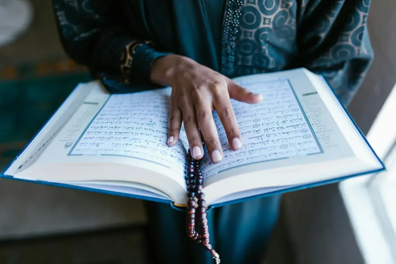 En hand vilar på en öppen Koran, med ett radband hängande över sidorna. En stilla stund för bön, kanske under fajr.