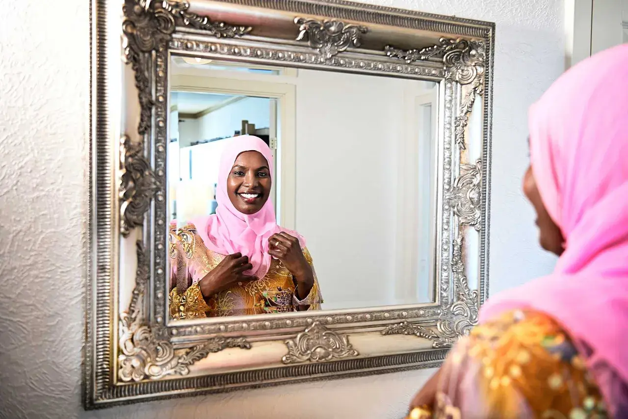 En kvinna i rosa hijab justerar sin klänning framför en utsmyckad spegel. Hon ler glatt, som om hon förbereder sig för ett speciellt tillfälle, kanske som Muhammeds fru Khadija en gång gjorde.