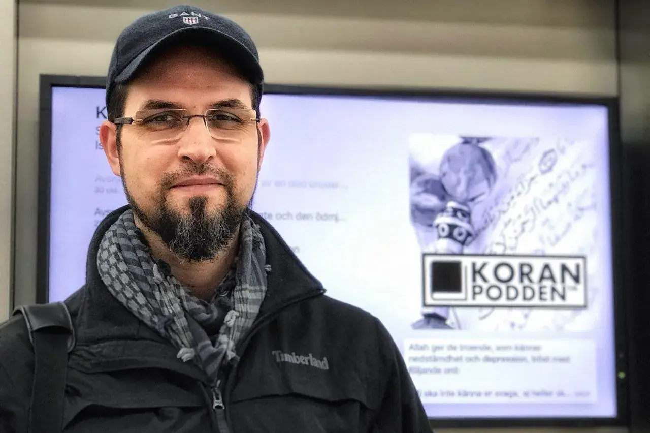 Man med skägg och keps ler mot kameran. I bakgrunden syns en skärm med texten "KORAN PODDEN" och arabisk skrift. Allahu alam.