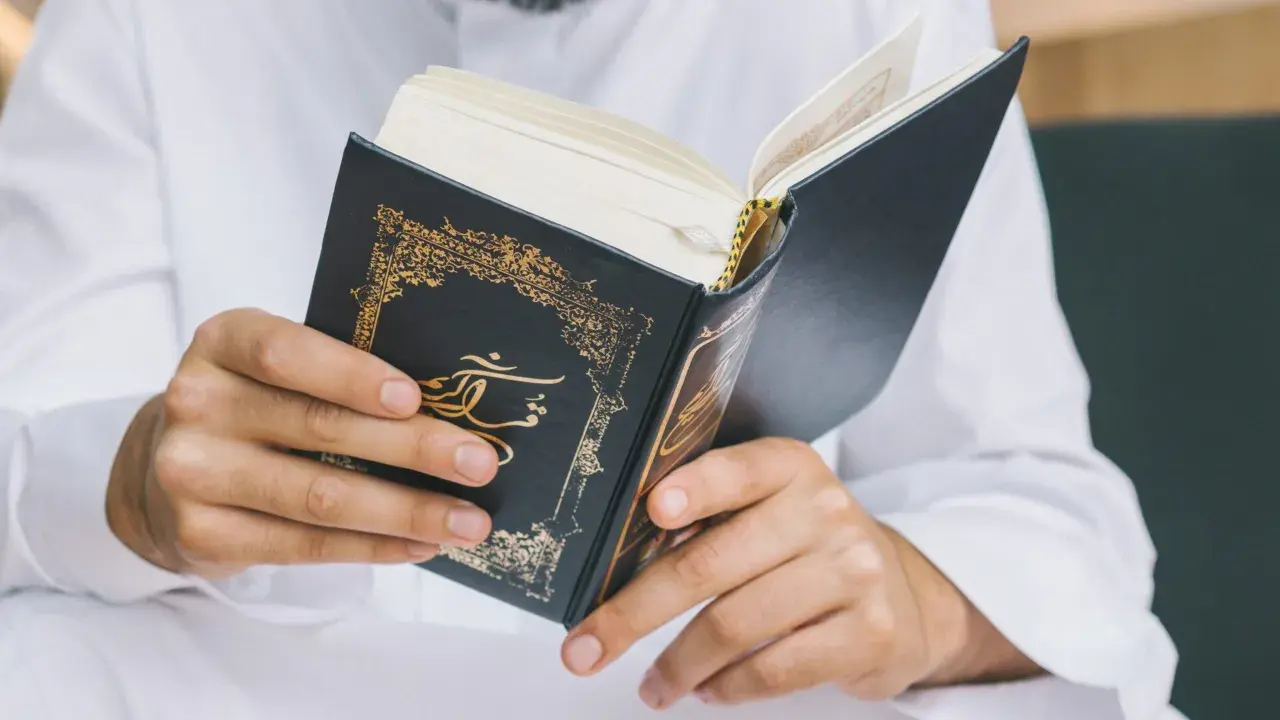 Man i vit klänning läser i en vackert dekorerad bok. Frågan om det är haram att raka skägget kan besvaras genom att studera religiösa texter.