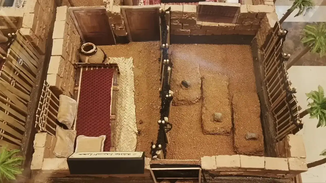 Modell av ett hus med en kruka, mattor och gravar. Kanske en scen från profeten Muhammeds tid.