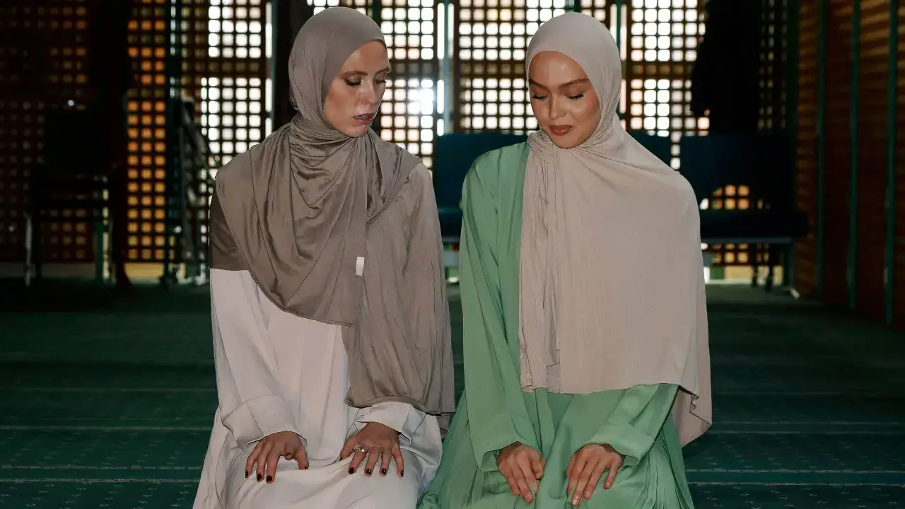 Två kvinnor i hijab knäböjer på en grön matta i en moské, förberedda för qiyam al layl.