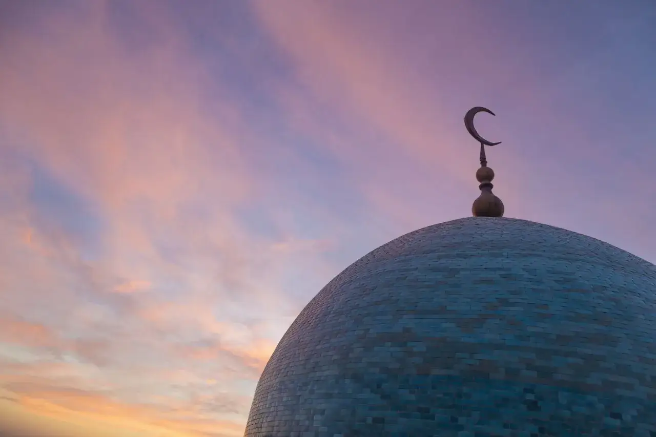 En blå kupol med en halvmåne islam-symbol mot en rosa och lila himmel.