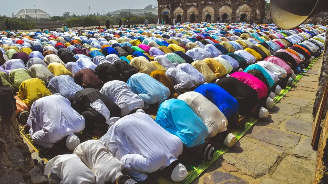 Stor grupp människor i färgglada kläder utför salat, en bön, på en stenlagd yta framför en gammal byggnad.